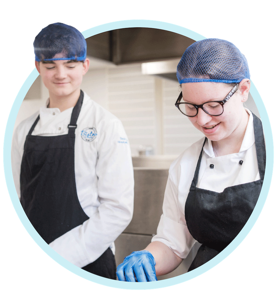Two catering students wearing hair nets, aprons and glove looking down preparing food in the kitchen
