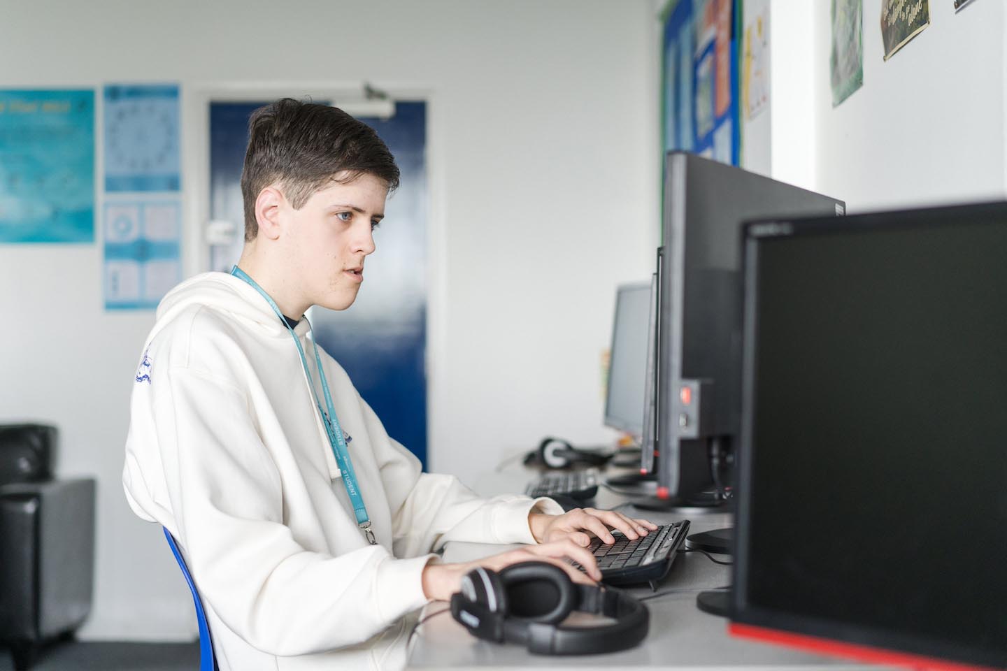 A young man wearing a white hoodie and a lanyard sits at a desk, focused on a computer monitor. His right hand rests on the keyboard while black headphones lie on the desk beside his left hand.