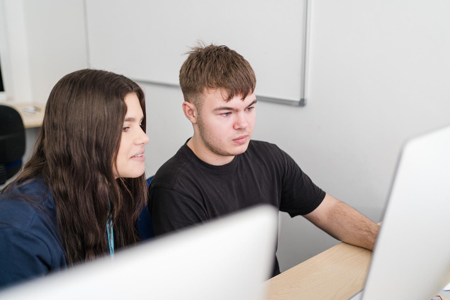 Two students sit side-by-side at a desk looking at a computer screen. On the left, a young woman with long dark hair leans in to look at the display while the young man beside her in a black t-shirt works with the mouse.