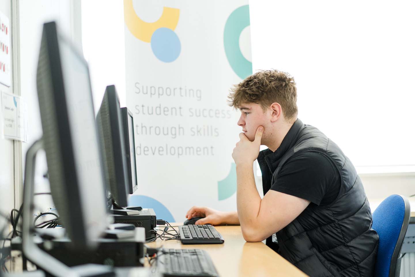 A side profile of a young man wearing a black vest and t-shirt sitting at a desk in a classroom or lab. He has one hand resting on his chin in thought while using a computer mouse with his other hand. In the background, a banner reads, "Supporting student success through skills development."