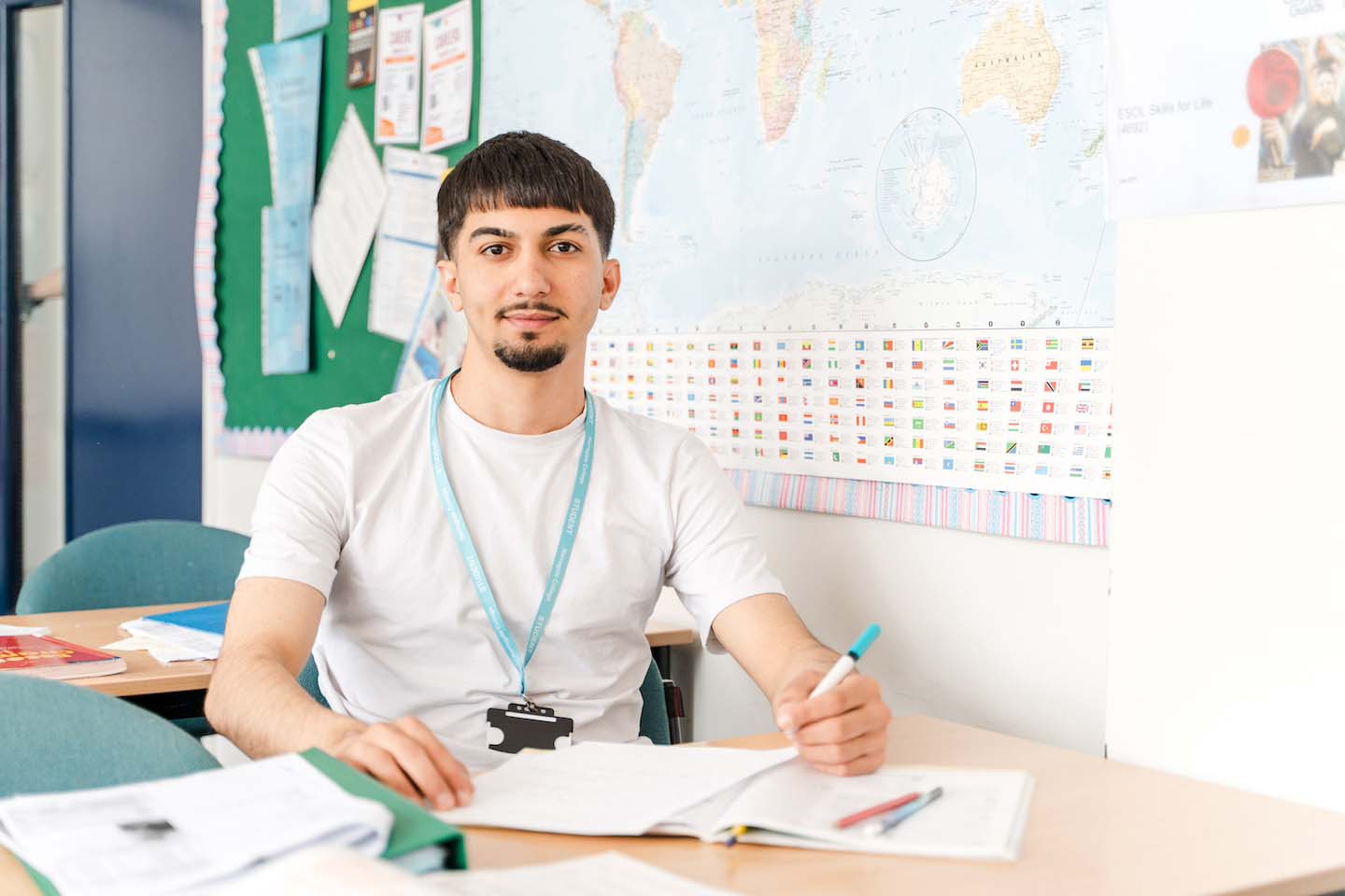 A young male student wearing a white t-shirt and a blue student lanyard sits at a desk in a classroom, holding a pen over a notebook. Behind him on the wall is a large world map with national flags.