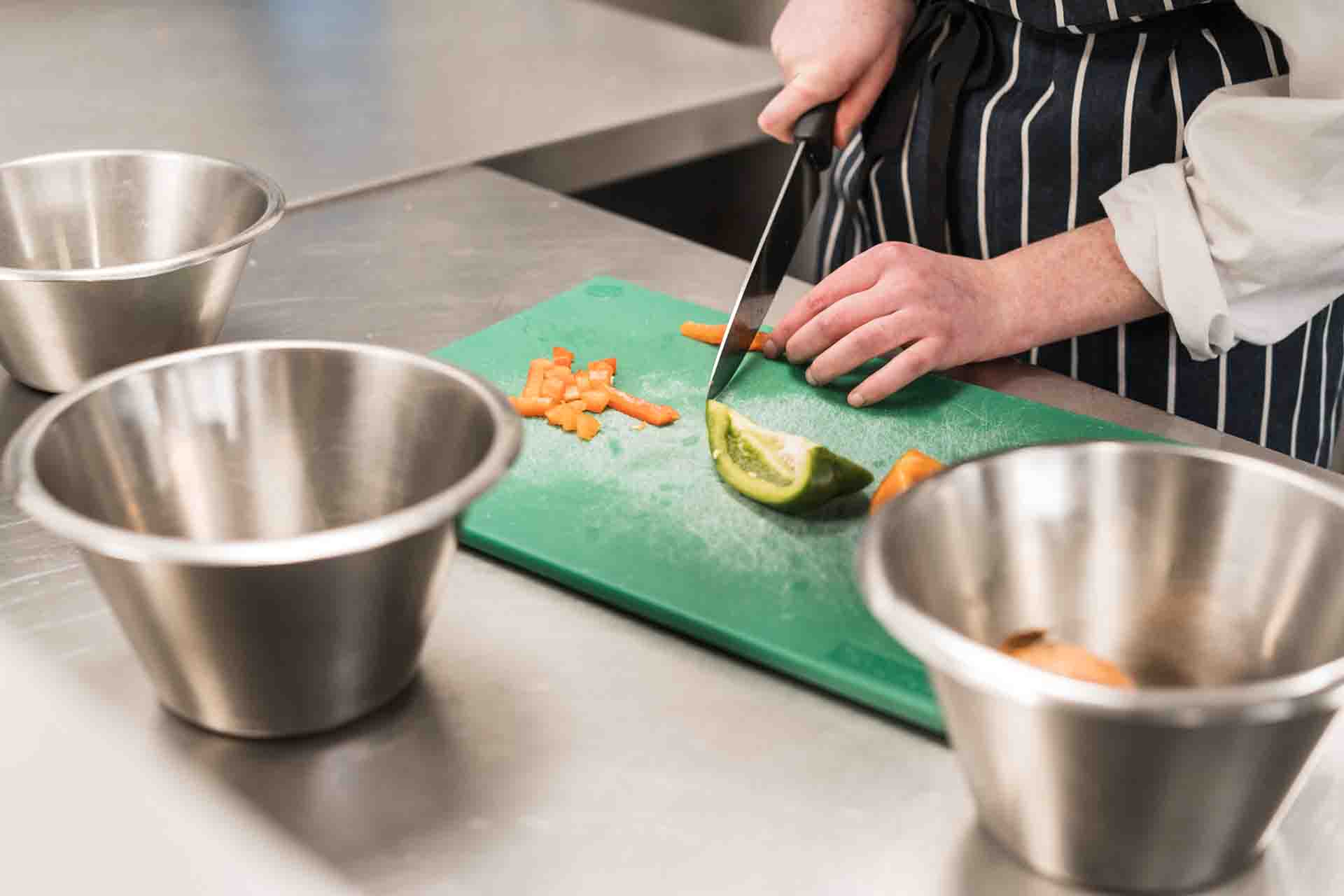 Student in black and white stripe apron chopping vegetables on a green chopping board with three stainless steel bowls on top of the preparing unit