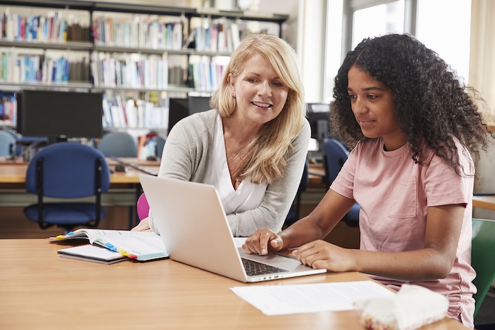 College student has individual tuition from teacher in library with laptop and notes on table
