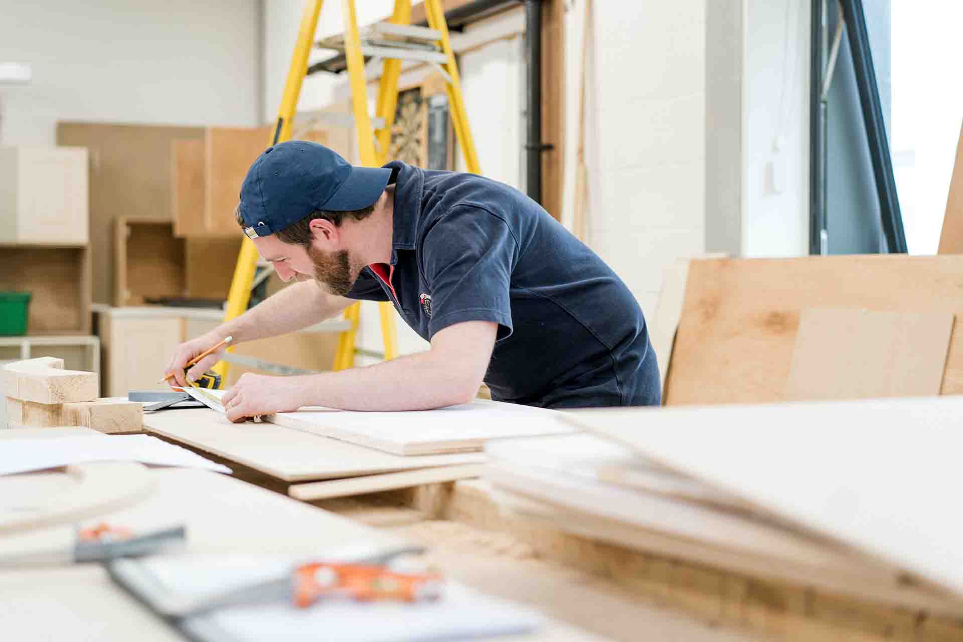 Student in blue polo shirt wearing a cap backwards, standing in the carpentry workshop with a measurement tape and pencil in hand taking some measurements of some wood planks