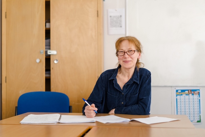 Teacher sitting down in a classroom holding a pen smiling