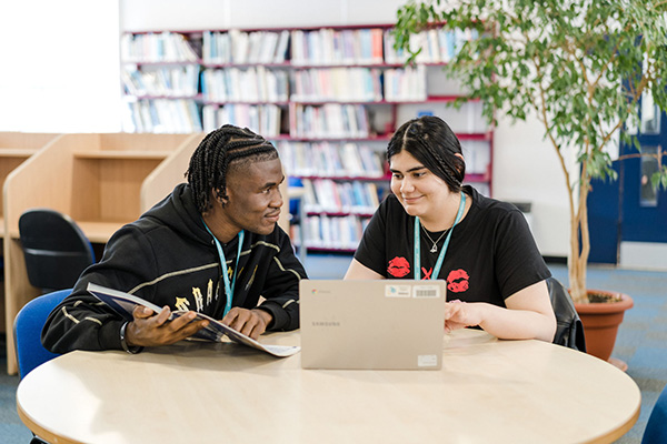 Two Harrogate College students working in the library working on a laptop and taking notes from a academic book